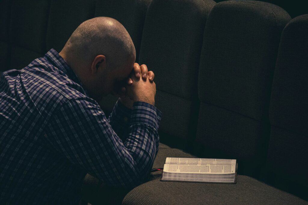 A man in a dimly lit room on his knees holding his hands together as an indication of prayer with an open bible. This is to show that we must be willing to change in order to have spiritual growth.