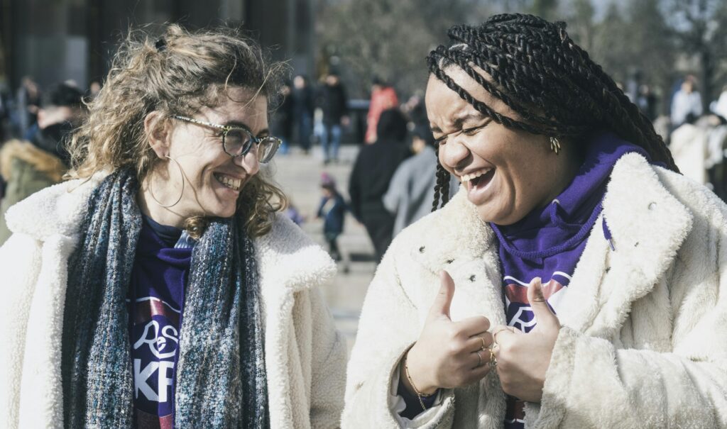 Two joyful women laughing together outdoors in winter coats, demonstrating authentic Christian fellowship and community connection in daily life