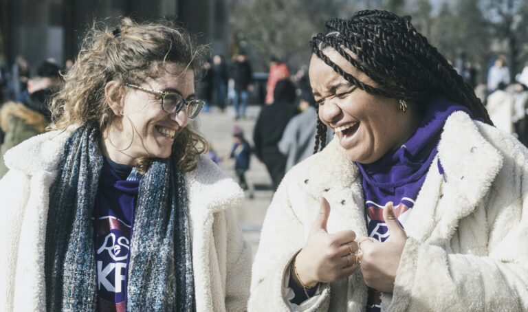 Two joyful women laughing together outdoors in winter coats, demonstrating authentic Christian fellowship and community connection in daily life