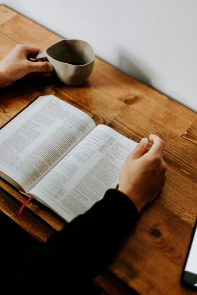 An open bible on top of a table with a woman reading it illustrating how to we should meditate on the scripture to hear God's voice