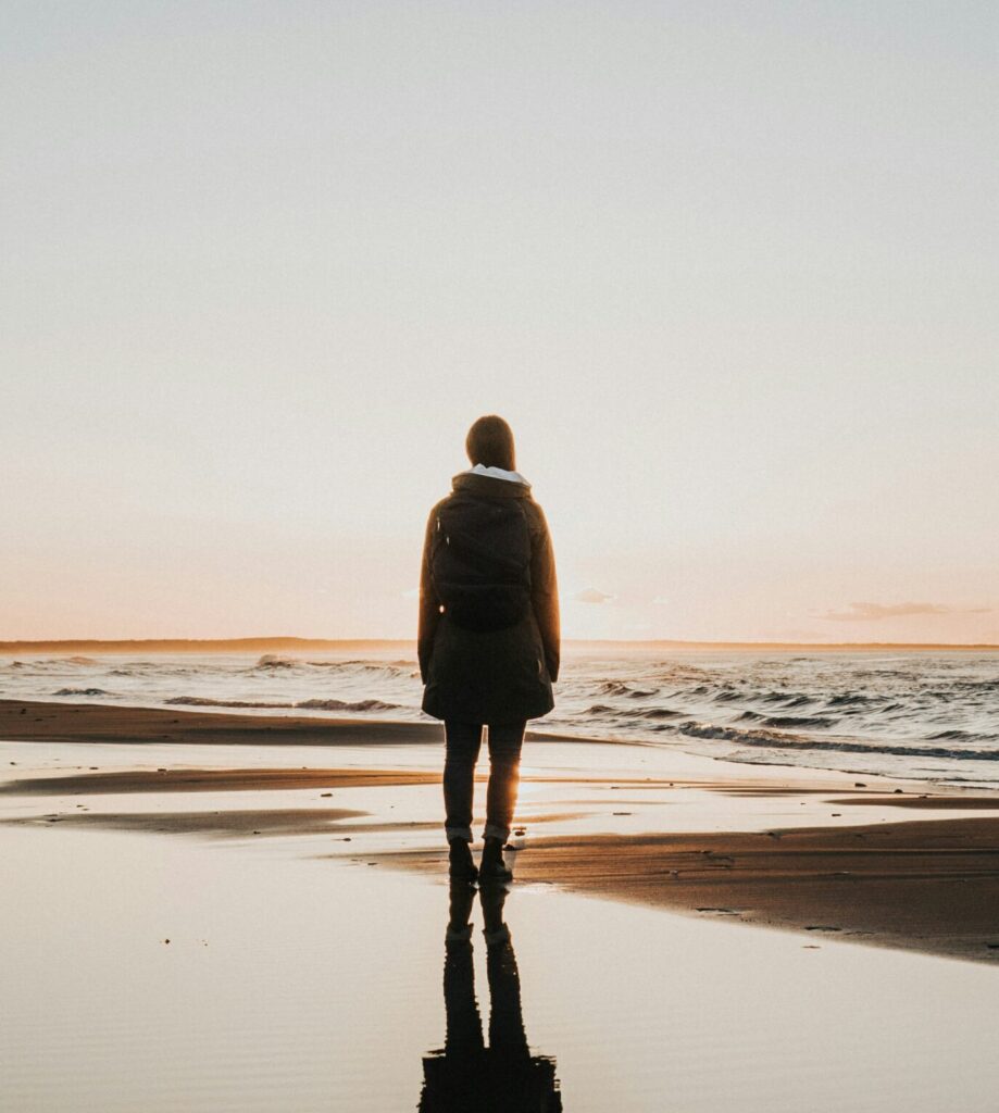 "Person standing alone on beach at sunset reflecting on water, symbolizing quiet meditation to hear God's voice"