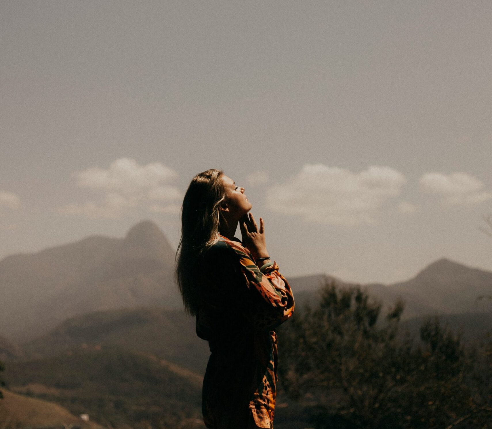 "Woman hearing God's voice through prayer on mountain at sunset with hands raised in worship"