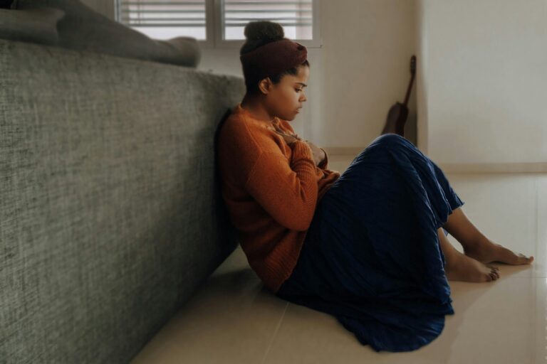 A young woman sitting on the floor with her hands on her chest, looking anxious and deep in thought, representing fear and seeking comfort from Bible verses.