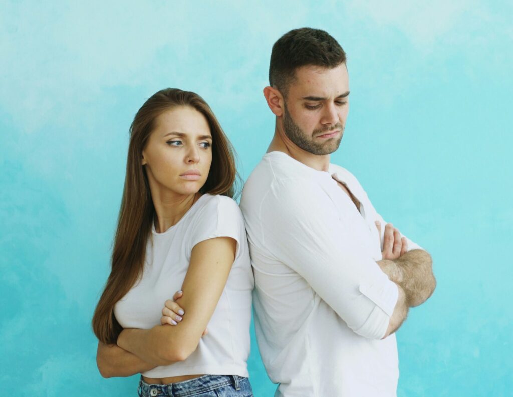 "Young couple standing back-to-back with arms crossed on turquoise background, displaying body language of disagreement and conflict in relationship"